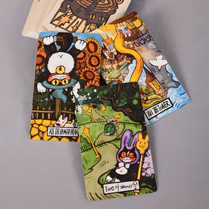 May include: A deck of tarot cards with whimsical illustrations. The cards feature colorful artwork of cats, rabbits, and other creatures in various scenes. The cards are labeled with titles such as "The Hanged Man" and "Five of Wands".