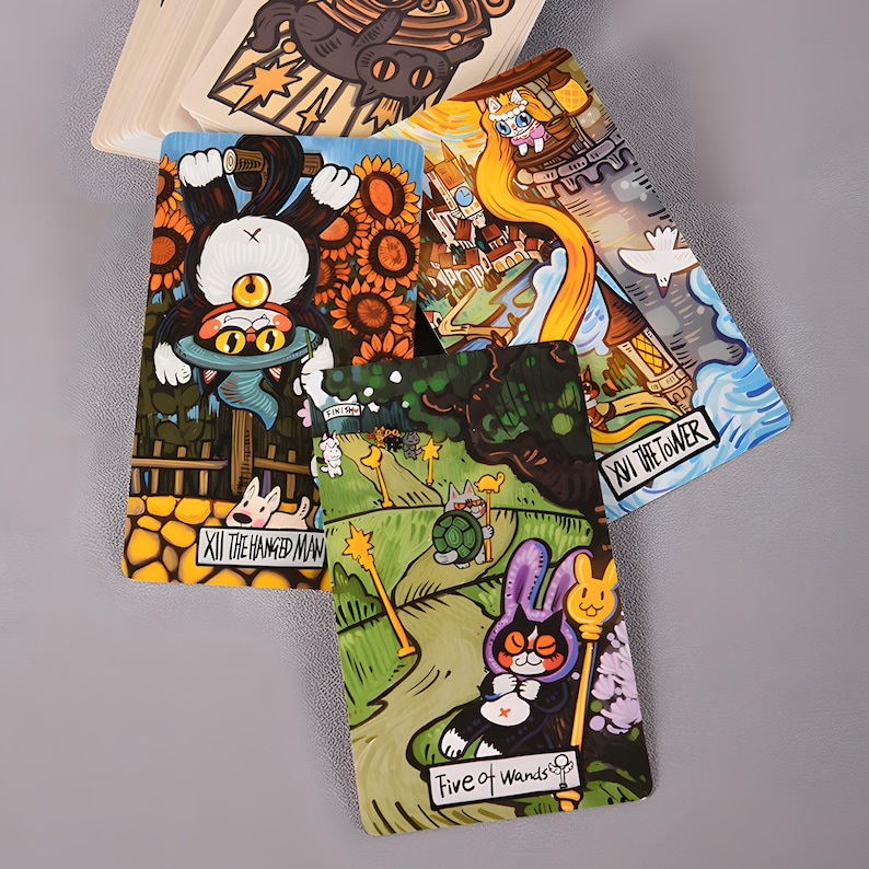 May include: A deck of tarot cards with whimsical illustrations. The cards feature colorful artwork of cats, rabbits, and other creatures in various scenes. The cards are labeled with titles such as "The Hanged Man" and "Five of Wands".