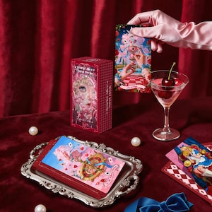 May include: A Scarlet Alice Tarot Deck box, a stack of tarot cards on a silver tray, and a single Ace of Cups card. The deck box features a character illustration and text. A martini glass with cherries and a blue ribbon are also present.