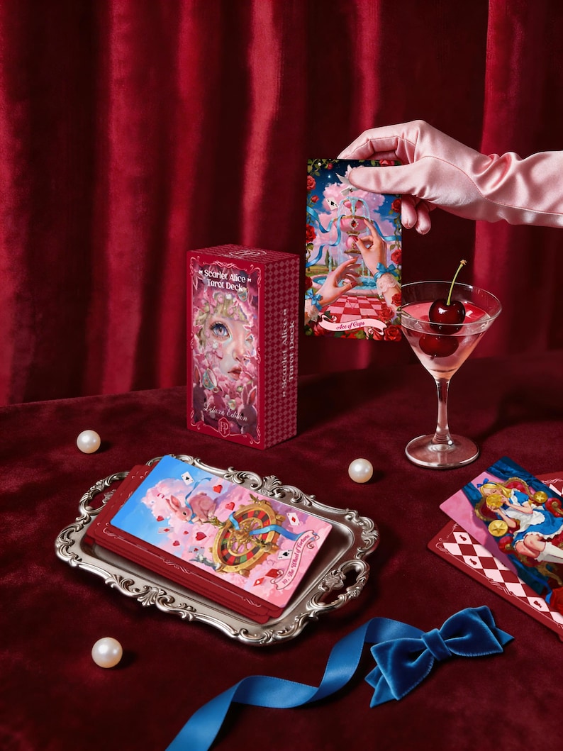 May include: A Scarlet Alice Tarot Deck box, a stack of tarot cards on a silver tray, and a single Ace of Cups card. The deck box features a character illustration and text. A martini glass with cherries and a blue ribbon are also present.