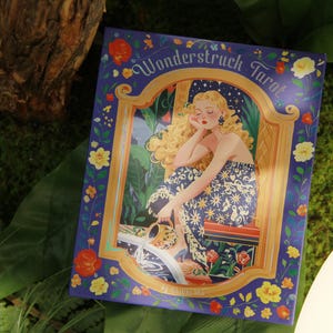 May include: A tarot card deck titled "Wonderstruck Tarot" with a woman in a floral dress. The card has a blue border with floral accents and gold trim. The background is green.