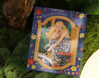 Könnte beinhalten: Ein Tarotkartenspiel mit dem Titel "Wonderstruck Tarot" mit einer Frau in einem Blumenkleid. Die Karte hat einen blauen Rand mit floralen Akzenten und goldenem Besatz. Der Hintergrund ist grün.