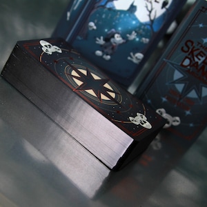 May include: A deck of cards with a dark navy blue background and a starburst design. The cards have a black edge and a red border. The deck is titled "The Skeleton Dance". The cards have a spooky theme.