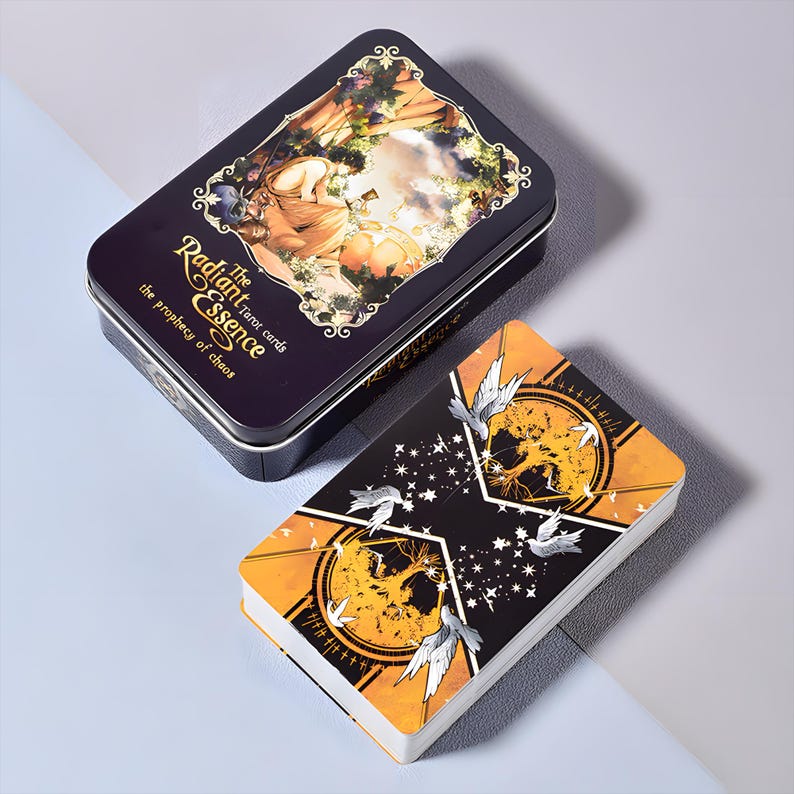 Puede incluir: Una baraja de cartas del tarot y su estuche a juego. El estuche es de color p&uacute;rpura oscuro con detalles dorados y el texto "The Radiant Essence tarot cards". Los dorsos de las cartas son negros y naranjas con un dise&ntilde;o celestial. La baraja est&aacute; abierta, revelando los bordes de las cartas.