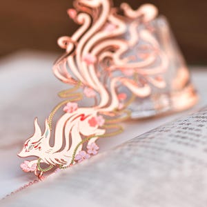 Könnte beinhalten: Kupferfarbener Metall-Lesezeichen mit einem stilisierten Fuchsmotiv und floralen Akzenten. Der Fuchs befindet sich auf einem offenen Buch, wobei sich der Schwanz des Lesezeichens nach oben erstreckt. Das Design weist filigrane Details und einen Hauch von Rosa auf.