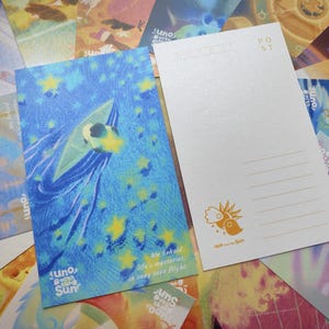 May include: Two postcards with illustrations. One features a blue background with a stylized figure and stars, with text that reads "Juno with the Sun". The other is white with lines for writing and a sun logo. Other postcards are in the background.