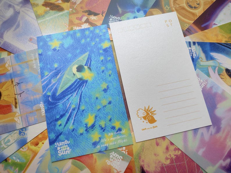 May include: Two postcards with illustrations. One features a blue background with a stylized figure and stars, with text that reads "Juno with the Sun". The other is white with lines for writing and a sun logo. Other postcards are in the background.