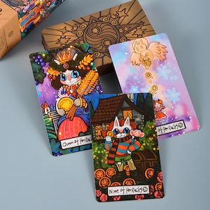 May include: A tarot card deck featuring whimsical illustrations. The cards depict anthropomorphic cats and other characters in vibrant colors. The wooden box has a yin-yang symbol and starburst design. The cards are labeled "Queen of Pentacles" and "Nine of Pentacles".