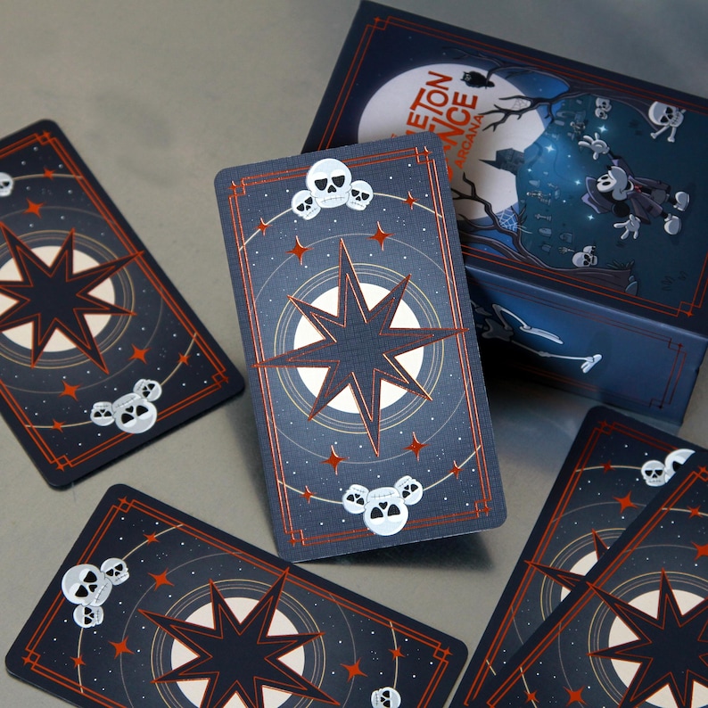 May include: A deck of tarot cards with a dark blue background, a starburst design, and skull accents. The cards are arranged around a matching box with the text "Skeleton Dance Arcana" and a spooky illustration.