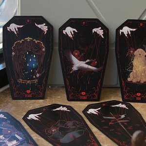 May include: A set of coffin-shaped cards with black backgrounds and red borders. Each card features a marionette-style illustration with white hands and strings. The cards display imagery including a mirror with stars, a stork, and a golden dog.