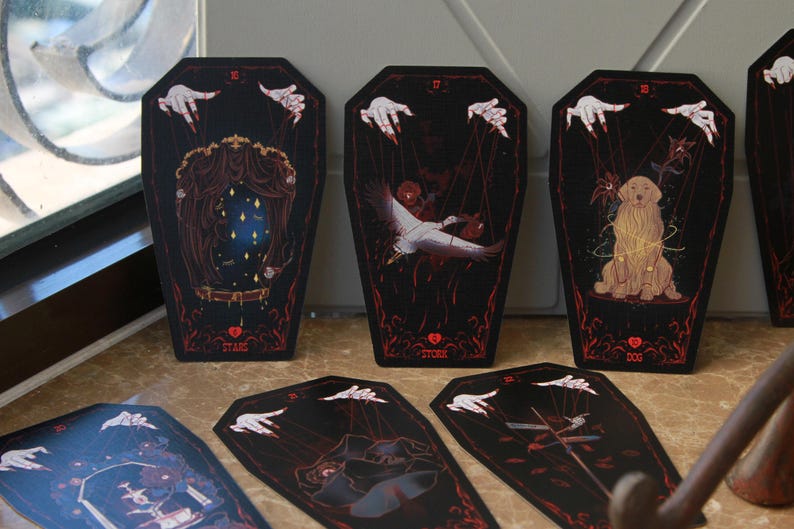 May include: A set of coffin-shaped cards with black backgrounds and red borders. Each card features a marionette-style illustration with white hands and strings. The cards display imagery including a mirror with stars, a stork, and a golden dog.
