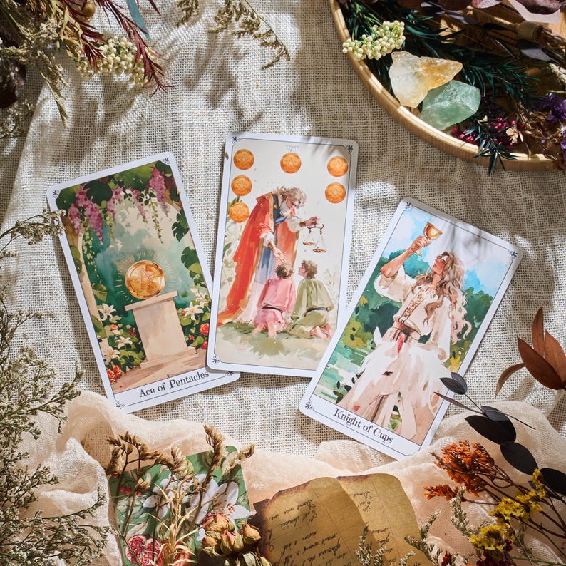 May include: Three tarot cards with watercolor illustrations are displayed on a beige surface. The cards are labeled "Ace of Pentacles" and "Knight of Cups." The third card depicts a figure with children. Dried flowers and crystals surround the cards.