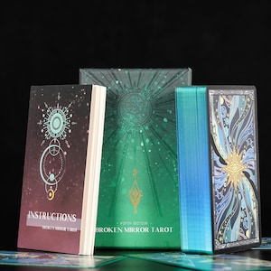 May include: A deck of Broken Mirror Tarot cards with a green box, an instruction booklet, and a card showing a blue and gold abstract design. The box features a sun-like emblem and the text Broken Mirror Tarot. The cards have metallic blue edges.