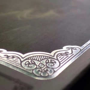 May include: A close-up of a silver-colored decorative border with an intricate design. The border is embossed with a floral pattern and has a raised edge.