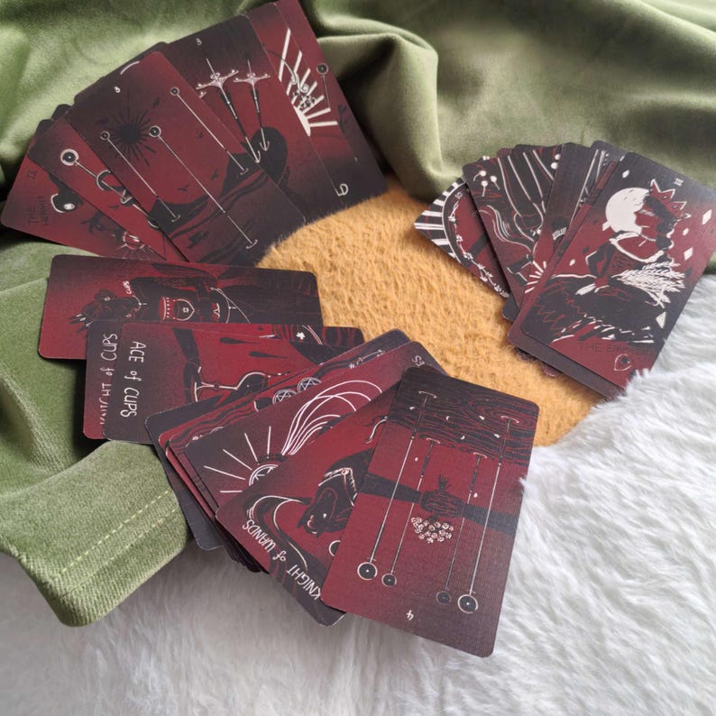Puede incluir: Una baraja de cartas de tarot con una combinaci&oacute;n de colores rojo oscuro y negro. Las cartas presentan ilustraciones y texto intrincados, incluyendo "Ace of Cups" y "Knight of Wands". Las cartas est&aacute;n abanicadas sobre una superficie texturizada.