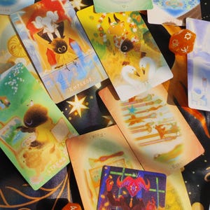 May include: A collection of tarot cards with whimsical illustrations and text, including "Emperor," "Empress," and "Strength." The cards are scattered on a dark surface, accompanied by orange dice.
