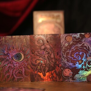 May include: Three rectangular tarot cards featuring intricate, colorful illustrations. The left card displays a large, detailed eye surrounded by tentacles. The center card shows a dark, monstrous figure. The right card depicts a swirling cosmic galaxy.