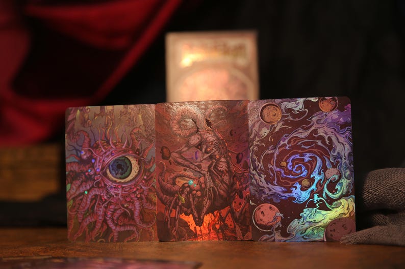 May include: Three rectangular tarot cards featuring intricate, colorful illustrations. The left card displays a large, detailed eye surrounded by tentacles. The center card shows a dark, monstrous figure. The right card depicts a swirling cosmic galaxy.