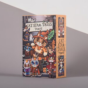 May include: A tarot card box featuring the title "CAT STAR TALES TAROT" with illustrations of cartoon cats in various costumes. The box has a light wood grain appearance and a colorful design with a whimsical theme.