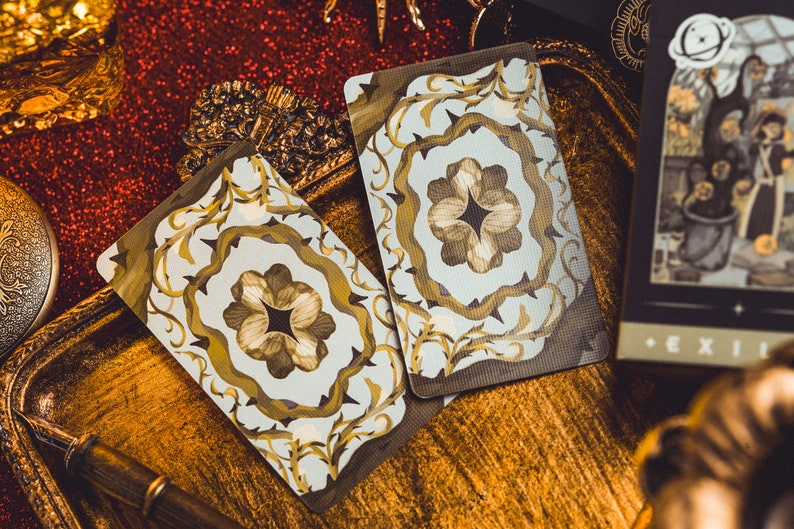 Op de afbeelding: Twee tarotkaarten met sierlijke bloemmotieven in goud en bruin, tegen een achtergrond van gouden en rode glitter. E&eacute;n kaart toont een gedetailleerde illustratie van een sc&egrave;ne met figuren en het woord "EXIT". De kaarten liggen op een gouden dienblad.