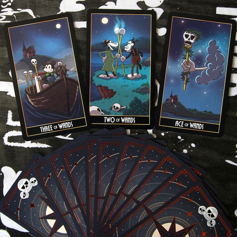 May include: A tarot card deck featuring illustrations of cartoon characters. The cards have a black border with gold trim and include the phrases "THREE OF WANDS", "TWO OF WANDS", and "ACE OF WANDS". The card backs are dark blue with a star and skull design.