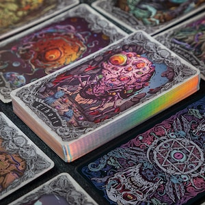 May include: A deck of rectangular playing cards with holographic edges, featuring intricate, colorful illustrations of grotesque creatures and occult symbols. The top card displays a multi-eyed, textured entity with the text Eihort printed below it.