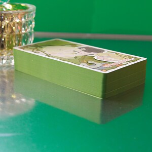 May include: A deck of tarot cards with green edges and a colorful illustration on the top card. The illustration features a figure in a natural setting with text. The cards are stacked on a reflective green surface.