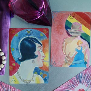 May include: Two rectangular tarot cards featuring number 29 and the letter A. One card shows a profile of a person with dark hair and a necklace. The other card depicts a person in a hat with a rainbow background and a small blue figure.