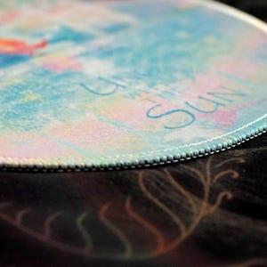 May include: Round mousepad with a colorful watercolor design in shades of blue, pink, and yellow. The edge is stitched with white thread. The words "Up with the Sun" are written in light blue.