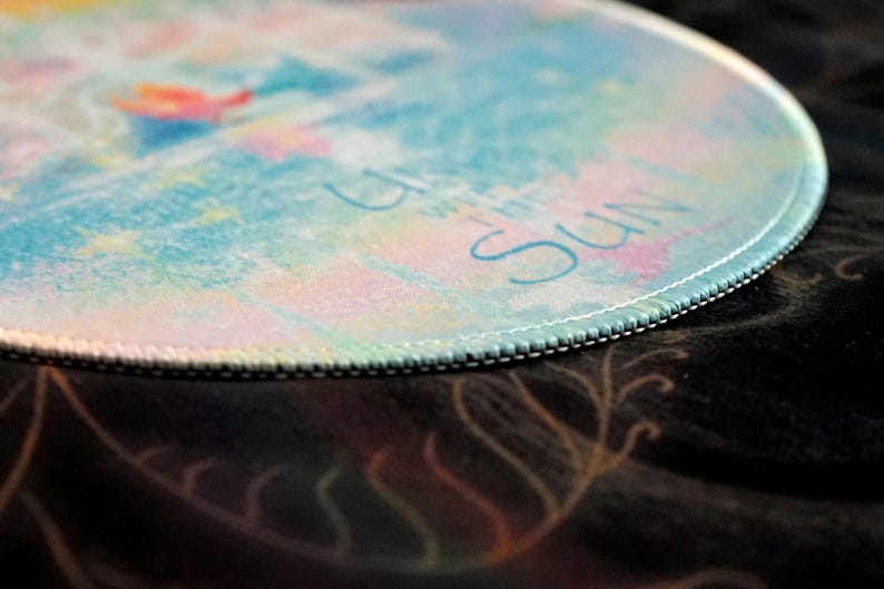 May include: Round mousepad with a colorful watercolor design in shades of blue, pink, and yellow. The edge is stitched with white thread. The words "Up with the Sun" are written in light blue.