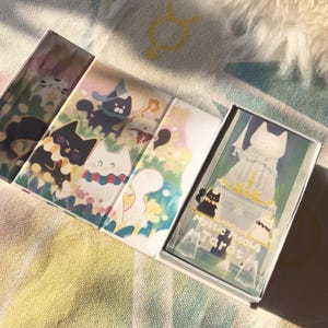 K&ouml;nnte beinhalten: Ein Tarotkartenspiel mit skurrilen Katzenillustrationen. Die Schachtel und die Karten zeigen farbenfrohe Kunstwerke von Katzen in verschiedenen Kost&uuml;men und Umgebungen. Das Deck tr&auml;gt den Titel "Mystic Meows Tarot" und enth&auml;lt ein Handbuch.