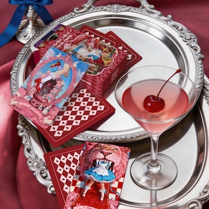 May include: A Scarlet Alice Tarot deck box and cards featuring illustrations of a character in a blue dress, a martini glass with a cherry, and a silver tray. The cards display the text Scarlet Alice Tarot Deck, The Fool, and Strength.