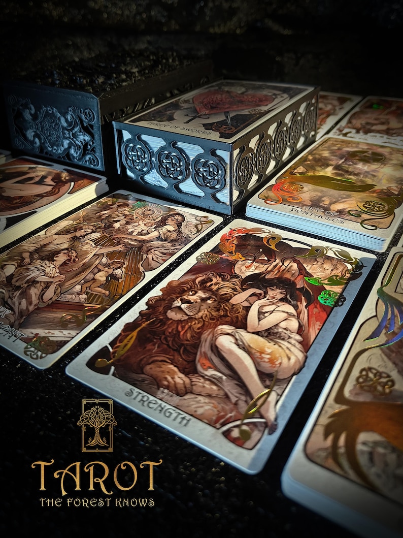 May include: A tarot card deck with decorative card boxes and individual cards. The cards feature detailed illustrations with gold accents. The word "STRENGTH" is visible on one card. The text "TAROT THE FOREST KNOWS" is at the bottom.