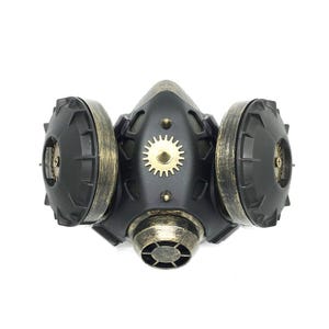 May include: A black and gold steampunk-style gas mask with two round, gear-shaped filters on the sides. The mask has a central, circular, gear-shaped design.