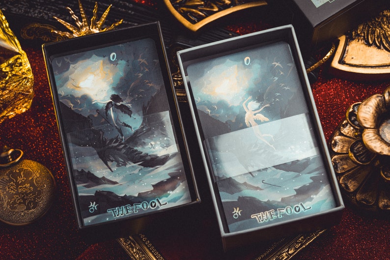 Op de afbeelding: Twee tarotkaartendecks in open dozen. De kaarten bevatten illustraties van een figuur met vleugels tegen een achtergrond van een maanverlichte lucht en oceaan. De tekst "THE FOOL" is zichtbaar op de kaarten. De dozen staan op een rood glitteroppervlak.