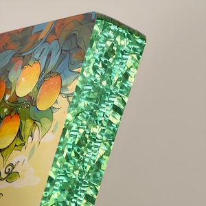May include: A stack of art prints with a vibrant illustration of orange fruit on a tree. The edges of the prints have a shimmering green effect, creating a unique visual texture. The artwork features a colorful, detailed design.