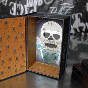 May include: A decorative box with a Halloween theme. The interior features a cartoon skull illustration against a graveyard backdrop. The box's interior walls are orange with a repeating skeleton pattern. The box is open, revealing the artwork.