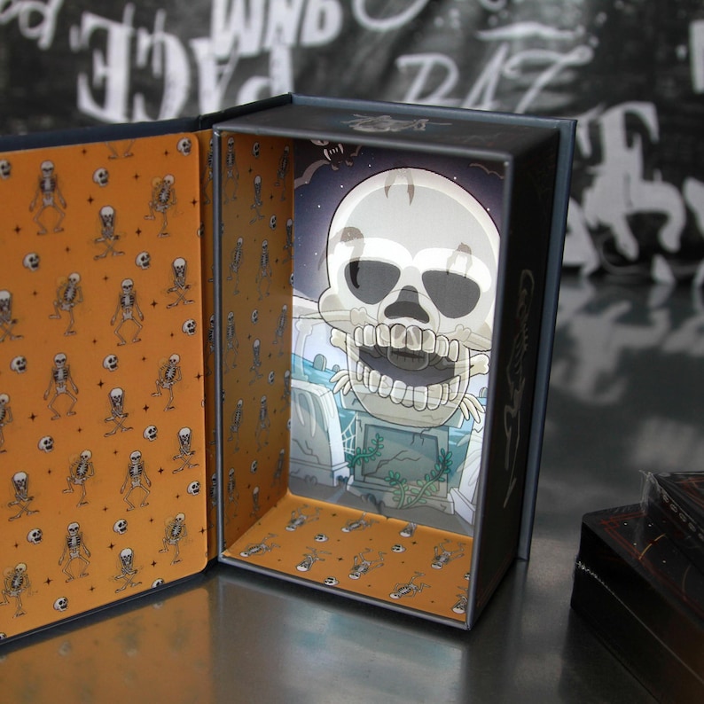 May include: A decorative box with a Halloween theme. The interior features a cartoon skull illustration against a graveyard backdrop. The box's interior walls are orange with a repeating skeleton pattern. The box is open, revealing the artwork.