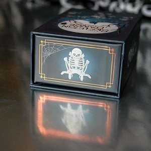 May include: A dark blue box with a silver skeleton design and the words "The Skeleton Dance" in white. The box has a gold border and a spiderweb design. The box is sitting on a reflective surface.