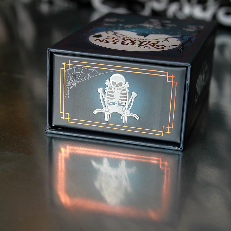 May include: A dark blue box with a silver skeleton design and the words "The Skeleton Dance" in white. The box has a gold border and a spiderweb design. The box is sitting on a reflective surface.