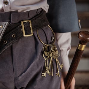 May include: A close-up of a black belt with a gold buckle and a decorative pattern. A large gold ring holds several antique-style keys. A wooden cane with a dark brown finish is in the background.
