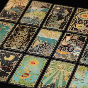 May include: A collection of rectangular tarot cards arranged in a grid, each featuring intricate, colourful illustrations with gold-toned line work. The designs depict various symbolic scenes, celestial bodies, and figures against dark backgrounds.