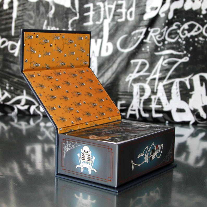 May include: A dark blue box with a hinged lid, featuring a Halloween theme. The box has a glowing skeleton design on the front and a spiderweb. The interior is orange with a pattern of skulls and spiders.