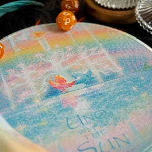 May include: Round, colorful mousepad with a watercolor design featuring two birds and the text "UNO WITH THE SUN." The background has a rainbow gradient. Two orange dice are visible.