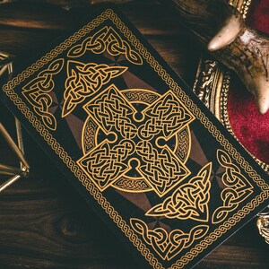以下が含まれることがあります： 金色のケルト結び目のデザインが施された黒いトランプ。カードは暗い背景に複雑な金色の模様が描かれている。