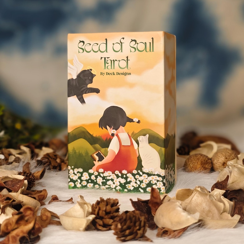 May include: A tarot card box with the title "Seed of Soul Tarot" and artwork featuring a girl, a white cat, and a black cat with wings. The background shows a sunset over green hills and a field of white flowers.