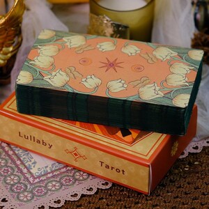 K&ouml;nnte beinhalten: Ein Tarotkartenspiel mit einem orangefarbenen Cover mit einem Blumendesign und einem Stern. Das Deck ist auf einer Schachtel mit den Worten "Lullaby Tarot" aufgedruckt. Das Deck hat gr&uuml;ne Kanten.