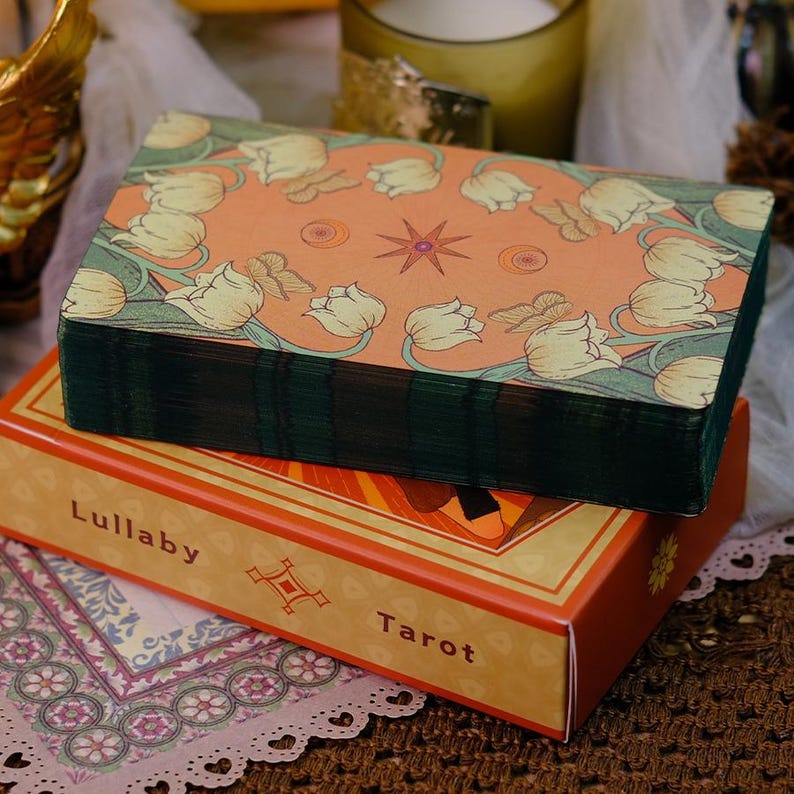K&ouml;nnte beinhalten: Ein Tarotkartenspiel mit einem orangefarbenen Cover mit einem Blumendesign und einem Stern. Das Deck ist auf einer Schachtel mit den Worten "Lullaby Tarot" aufgedruckt. Das Deck hat gr&uuml;ne Kanten.