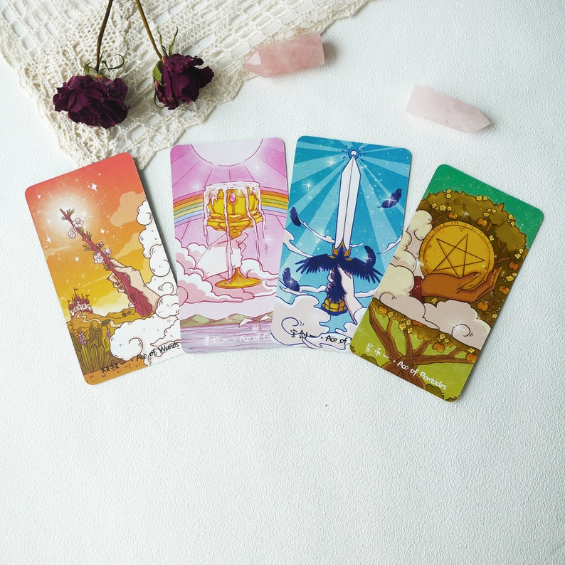 May include: Four tarot cards with colorful illustrations and gold accents. The cards are from the suit of wands, cups, swords, and pentacles. The cards are laid out on a white surface with pink crystals and a pink stone.