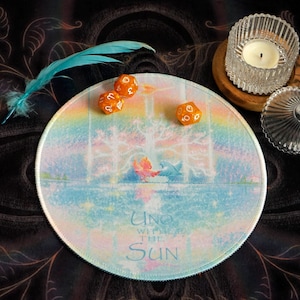 May include: A round, pastel-colored mousepad with the text "Uno with the Sun" and a whimsical illustration. Three orange dice and a blue feather are on the dark background. A glass candle holder and a small glass jar are also present.
